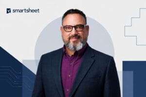 Portrait of Drew Garner, Smartsheet’s first Chief AI Officer, in a navy blazer and purple shirt against a subtle geometric corporate background with the Smartsheet logo.